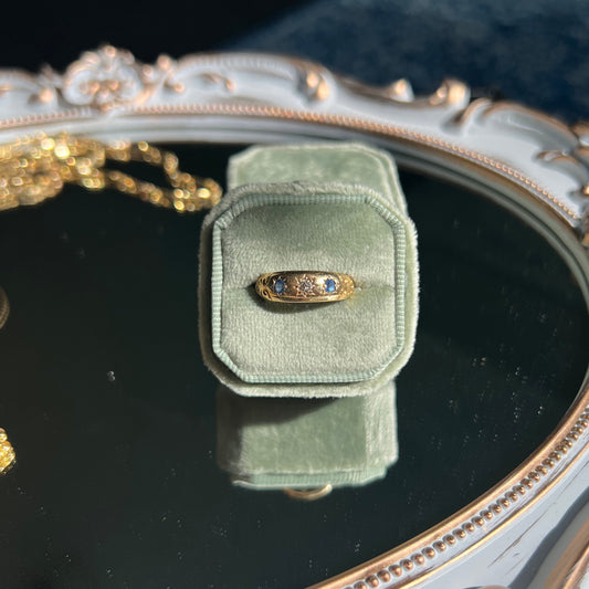 yellow gold band ring with sapphires and diamond in a light green ring box on top of a mirror